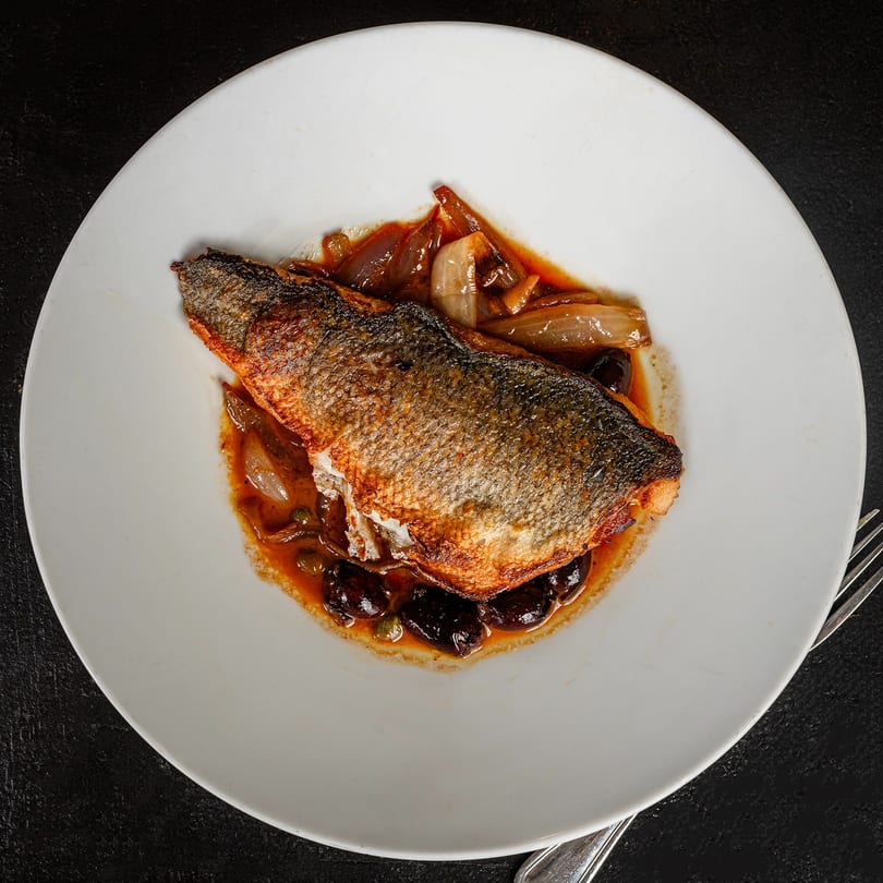 Pan-seared fish with crispy skin, served on a white plate with onions, olives, and capers in sauce.